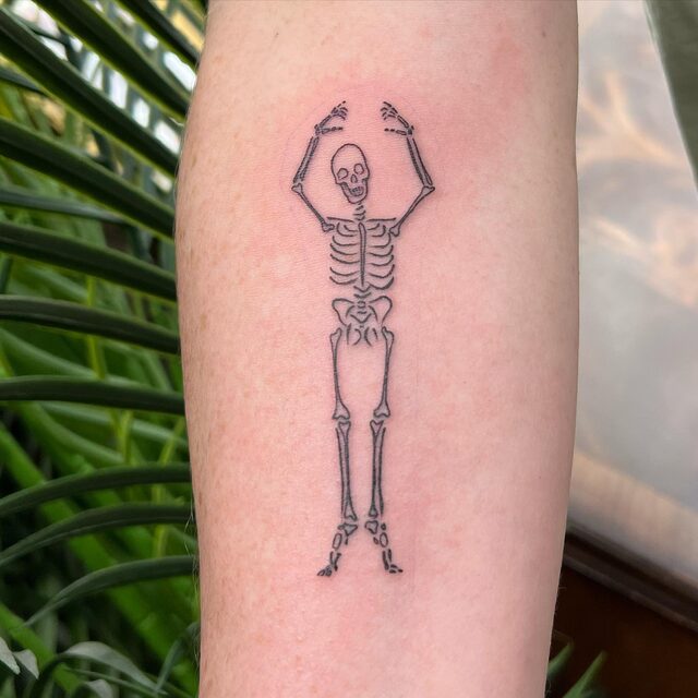 Fine-line tattoo of a dancing skeleton with its arms raised, on a forearm