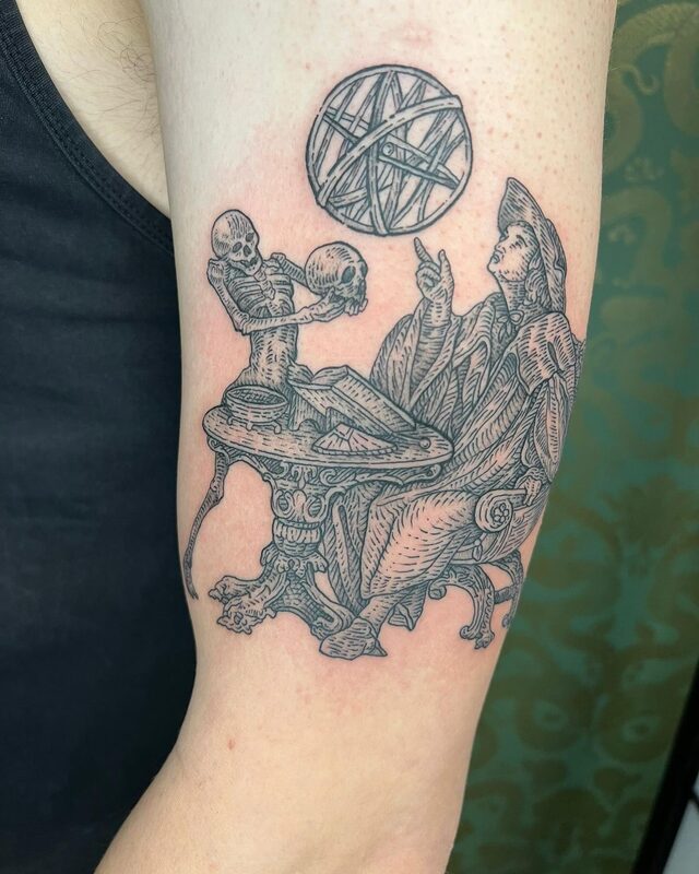 Etched tattoo of a skeleton showing a skull to a robed scholar at a table beneath an armillary sphere, on an upper arm