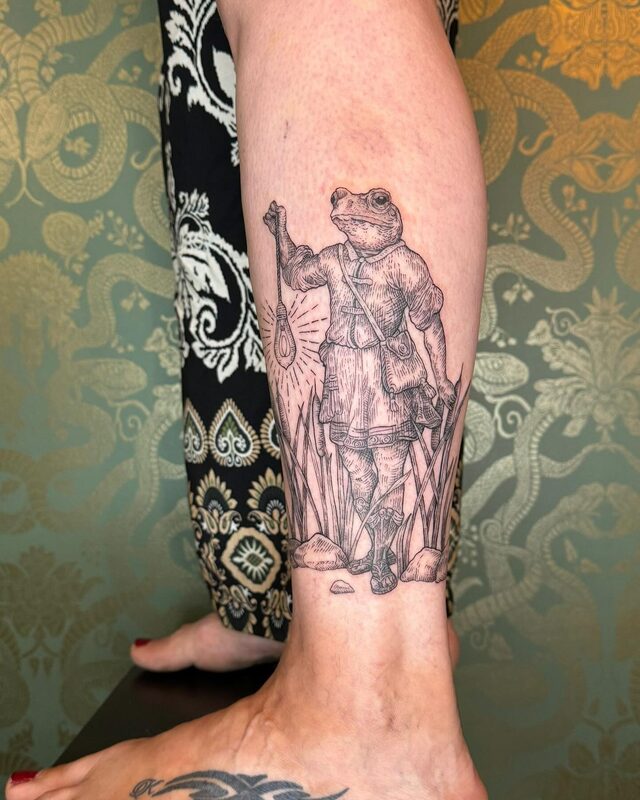 Fine-line etching of a frog in a traveler’s cloak holding up a lantern, tattooed on a lower leg