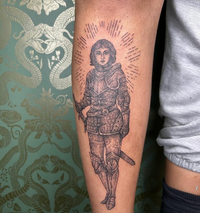 Etched tattoo of Joan of Arc in plate armor holding a sword, ringed by radiating lines, on a forearm