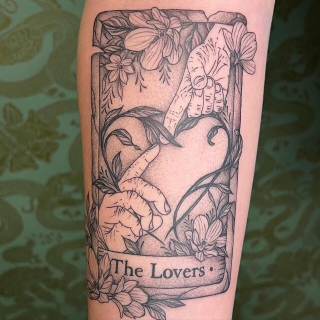 Tarot card tattoo titled The Lovers, two hands reaching toward each other inside a heart of vines and flowers, on a forearm