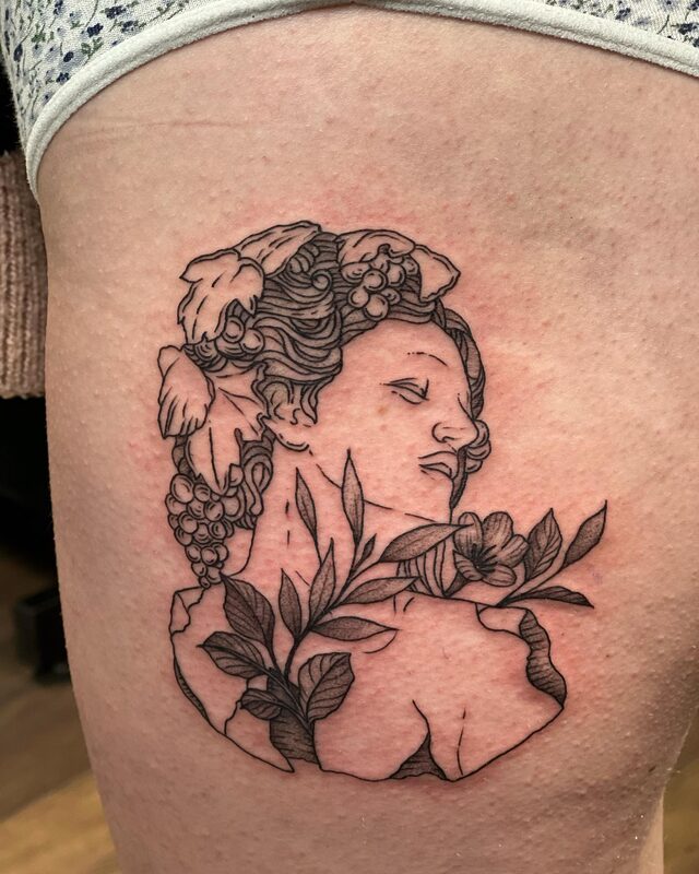 Linework tattoo of a classical statue’s head with grapes in her hair, surrounded by leaves and flowers, on a thigh