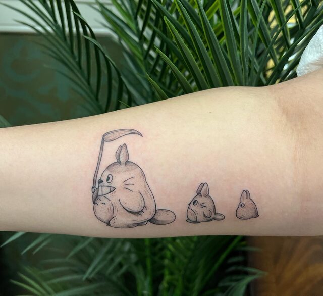 Fine-line tattoo of Totoro holding a leaf over his head, followed by two small Totoros, on a forearm