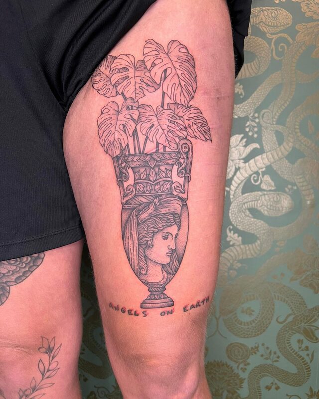 Etched tattoo of a classical vase with a woman’s profile and monstera leaves growing out of it, above the words Angels on Earth, on a thigh